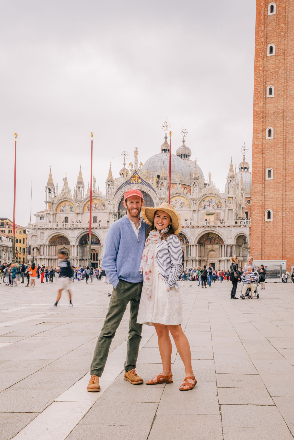 A smiling couple standing in Saint Mark's Square in front of the golden domes and mosaic arches of the basilica on a cloudy day