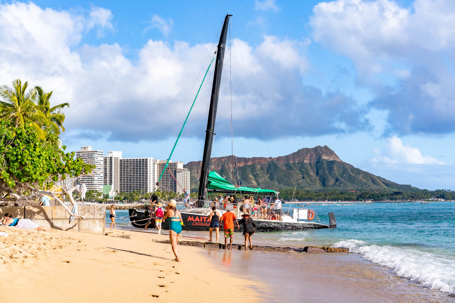 Sailing catamaran pulled up on Waikiki Beach with passengers gathered on the sand and Diamond Head rising in the distance