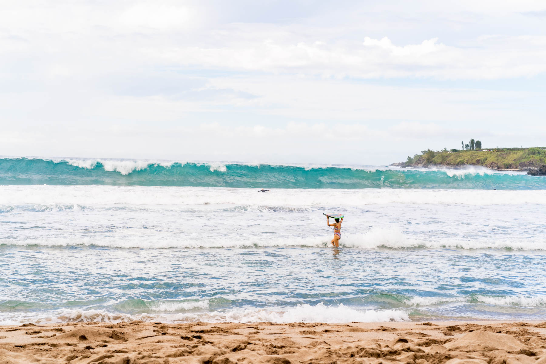 Woman carrying a surfboard on her head wading into shallow water as a wave breaks behind her on a Maui beach