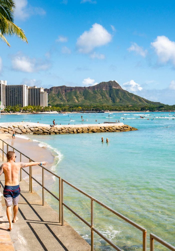 Couple walking a beachfront path along Waikiki with Diamond Head crater and turquoise water in the background under a clear sky