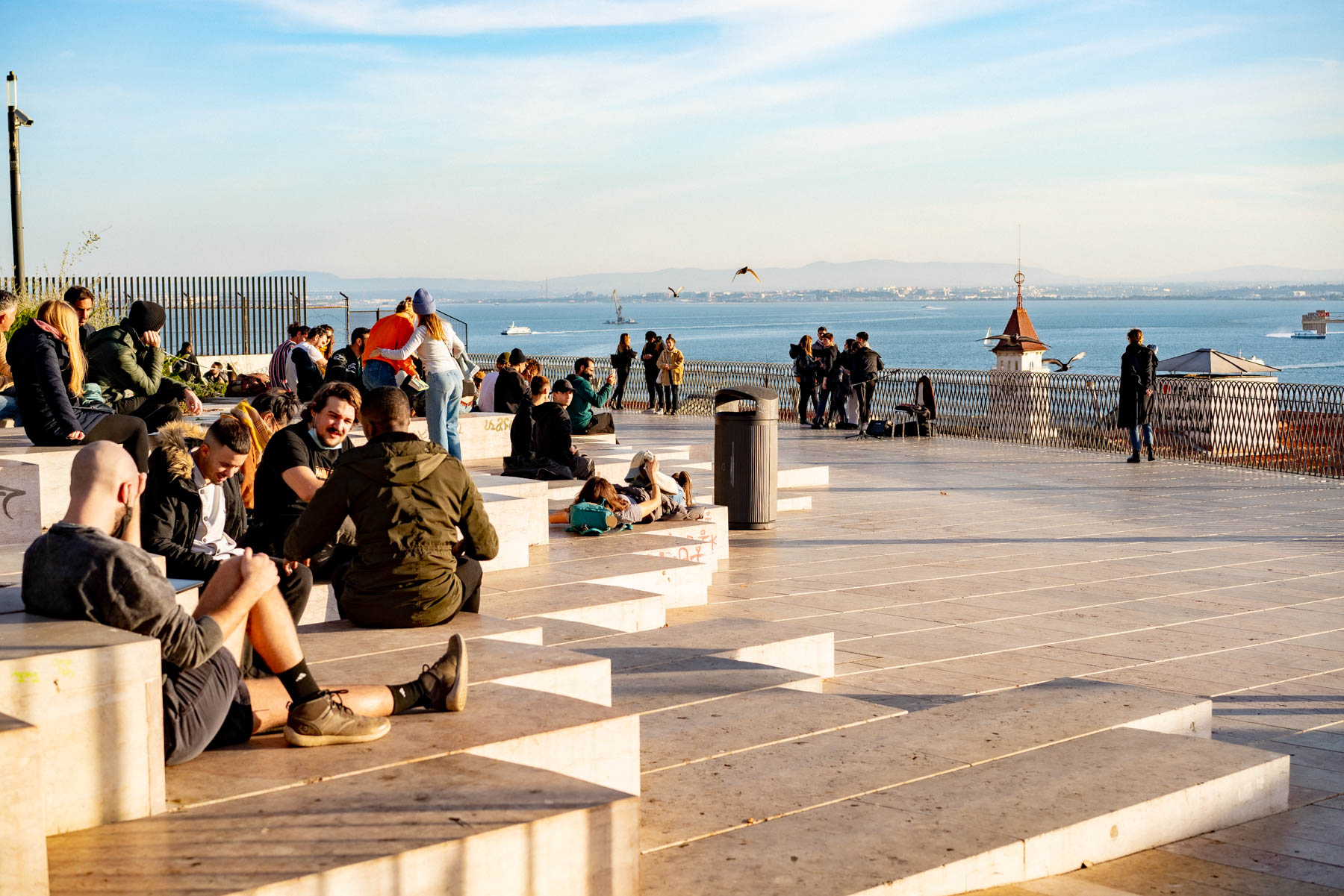 People in coats and beanies sitting on wide stone steps at a Lisbon viewpoint terrace looking out over the Tagus river with a boat on the water