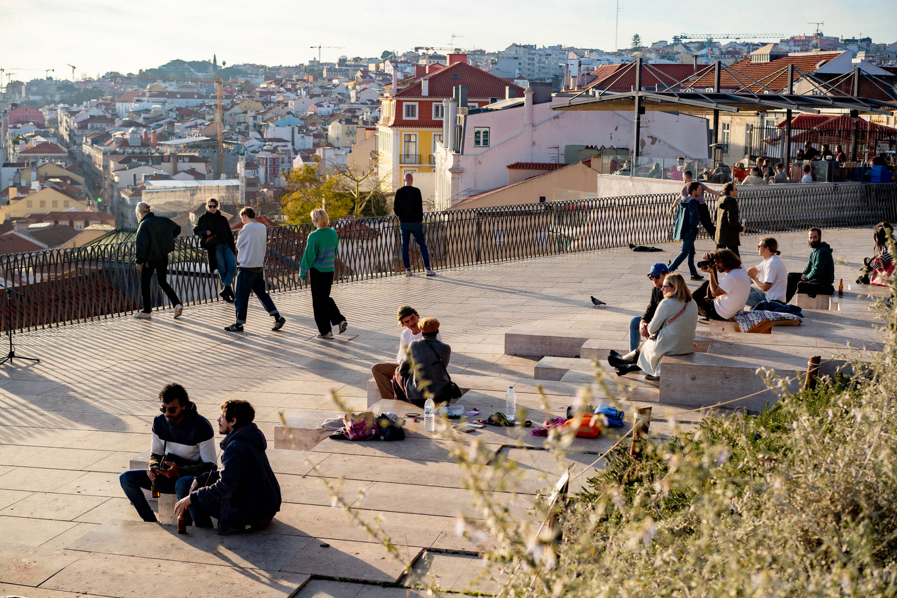 Low golden light across a stone viewpoint terrace above Lisbon's tiled rooftops, with people in jackets sitting on the steps and walking the railing