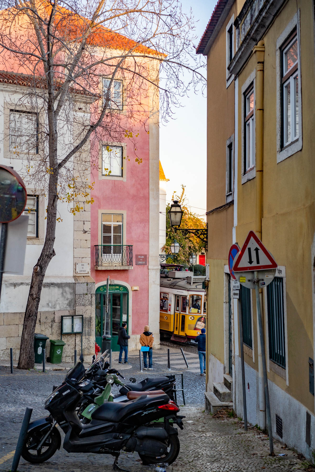 A narrow Alfama street with pink and yellow buildings and a leafless tree, a yellow tram waiting at the bottom of the hill and a few people in jackets standing nearby