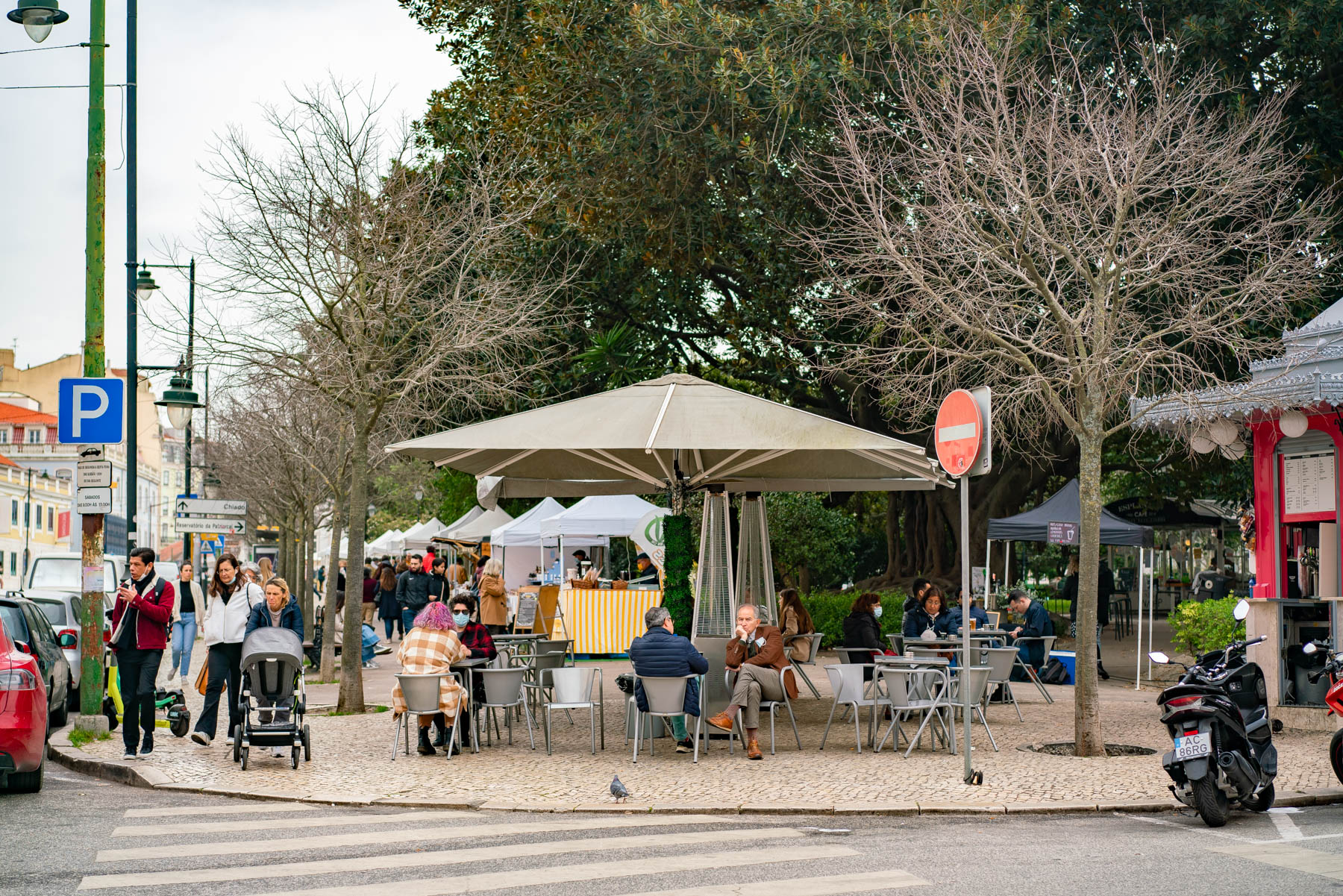 A kiosk cafe in a Lisbon garden square under bare plane trees, with people in coats and scarves sitting at metal tables and market stalls set up behind them