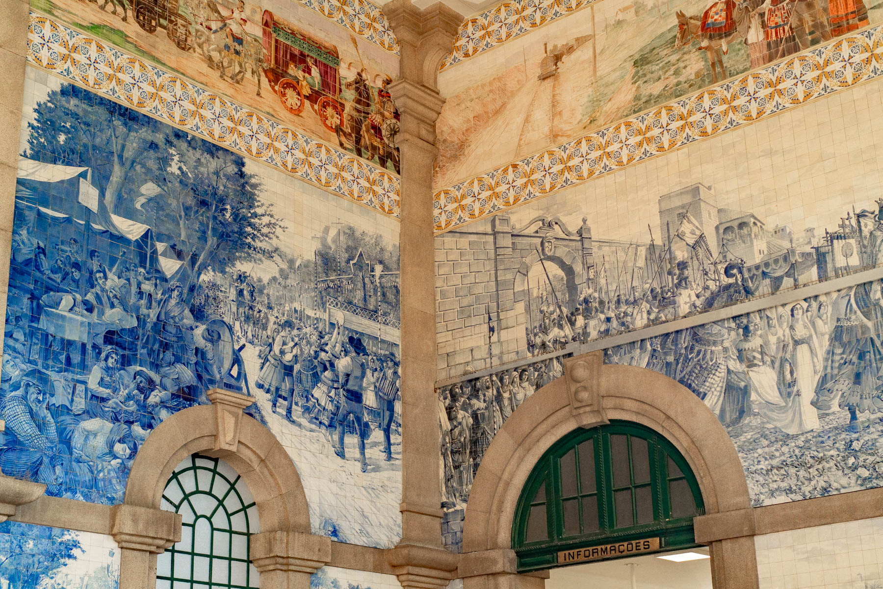 The tiled entrance hall of Sao Bento station in Porto, its walls covered floor to ceiling in blue and white azulejo panels of processions and market scenes above a green-framed information window