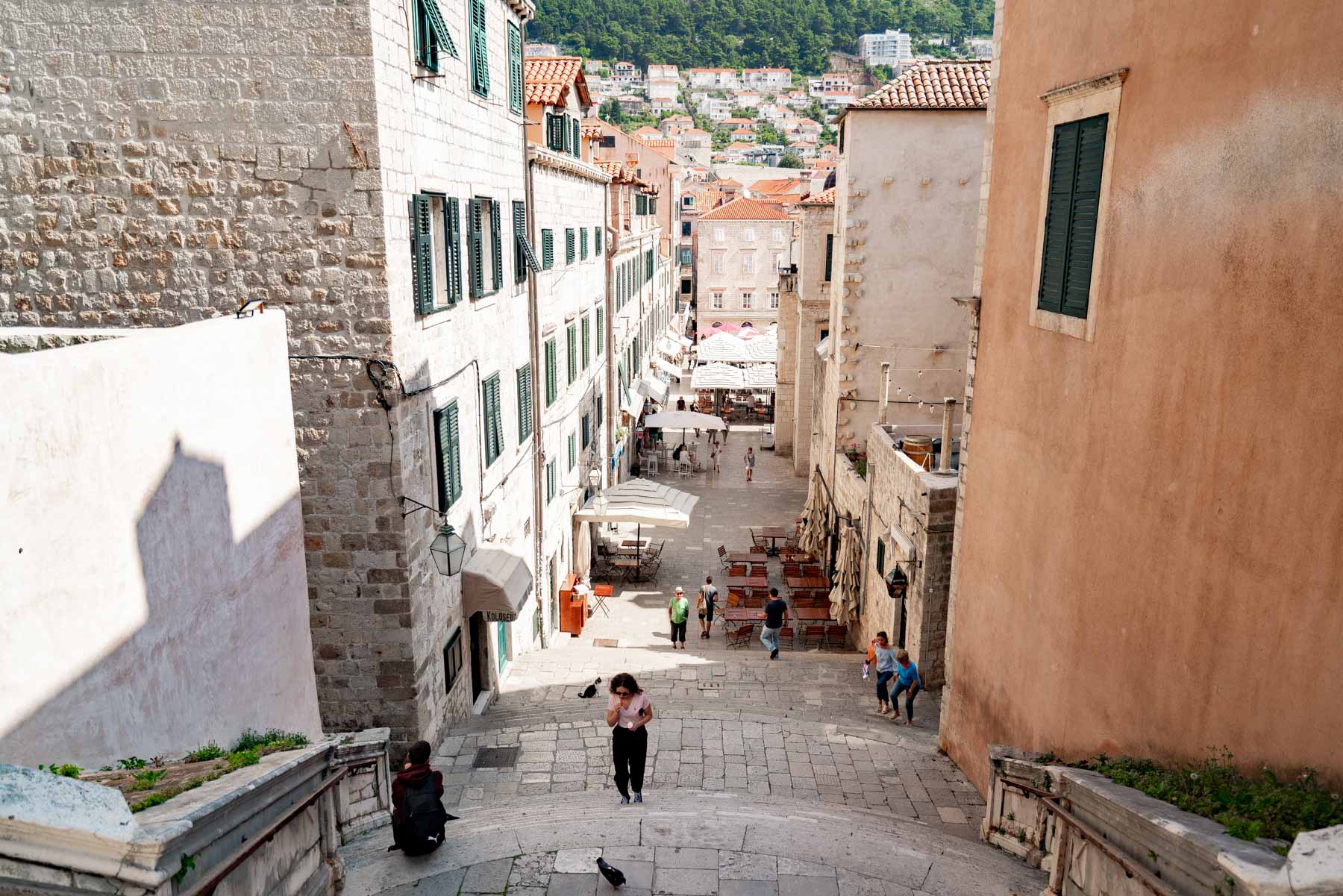 Looking down a wide staircase in Dubrovnik toward a quiet lane of cafe umbrellas and pale limestone buildings