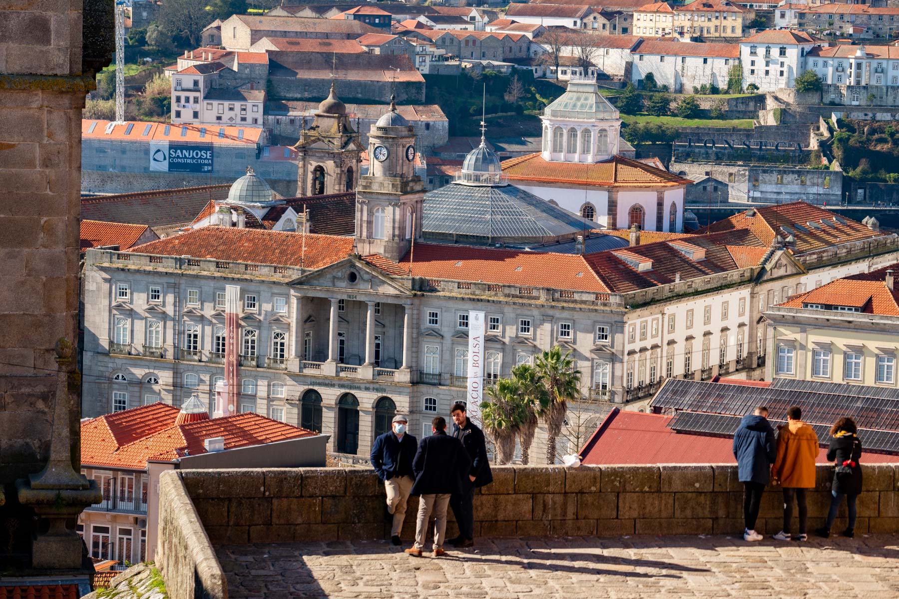 A view from a stone terrace over Porto rooftops toward the domed glass roof and columned facade of the Palacio da Bolsa, with visitors in heavy winter coats leaning on the wall in the foreground