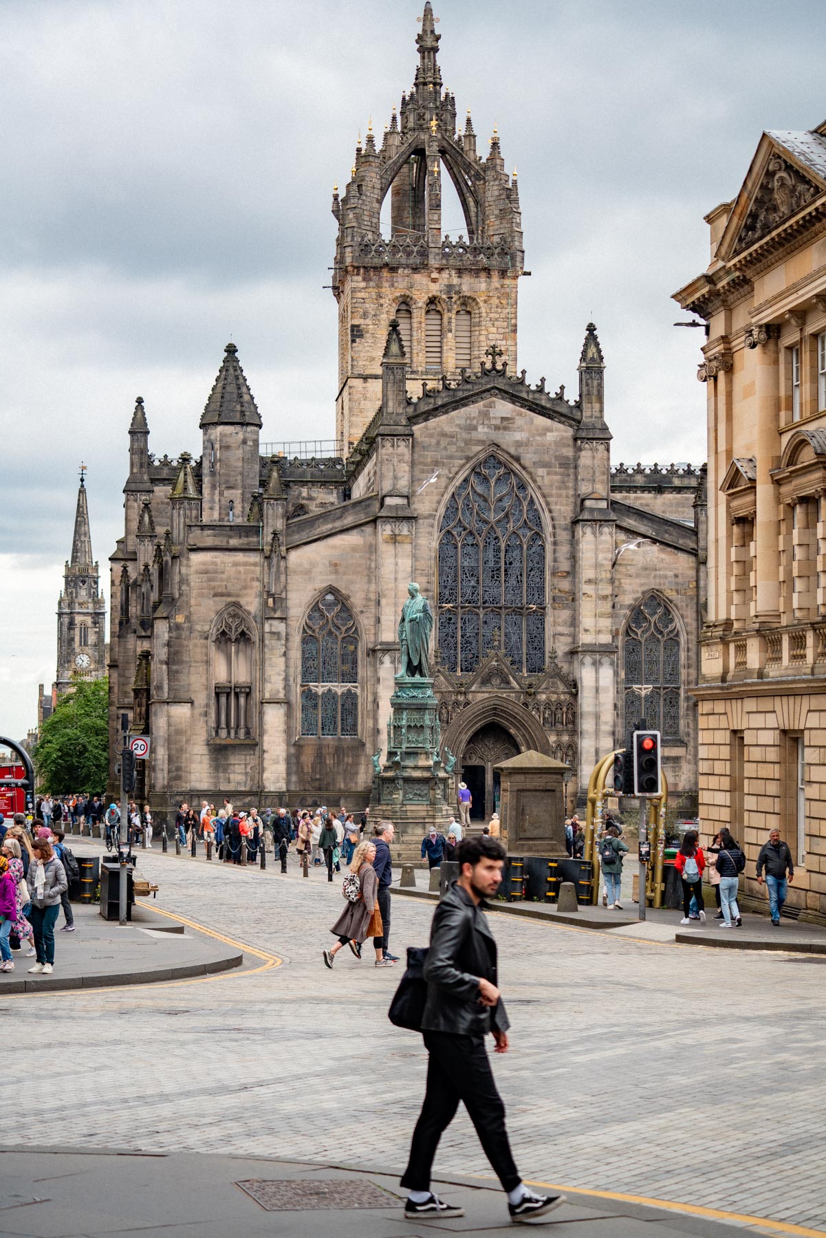 The tall stone crown spire of St Giles' Cathedral rising above people crossing the Royal Mile in Edinburgh under a cloudy sky